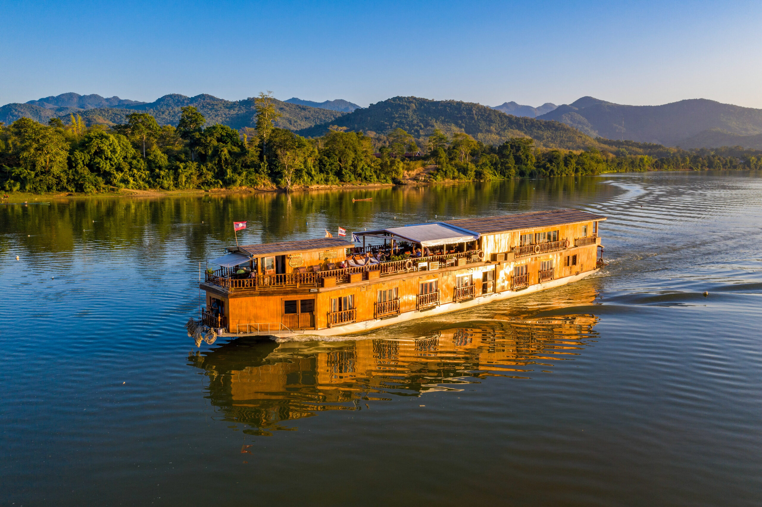 Wooden river cruise boat reflected in calm Mekong water at sunset, mountains behind